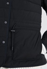 Svart puffjacka med quiltat mönster, utrustad med tryckknappar, dragsko vid nederkanten och en framficka. Slät textur.