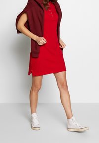 Czerwona sukienka bez rękawów z kołnierzykiem zapinanym na guziki, w połączeniu z bordowym dresowym swetrem z kapturem i białymi trampkami za kostkę. Gładka tkanina i casualowy styl.