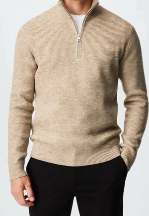 Pull en maille beige côtelée avec un col zippé à l’avant, porté sur une chemise blanche, associé à un pantalon noir, mains détendues le long du corps.