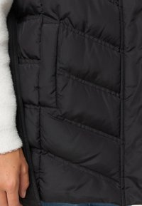 Mustat pehmustetut liivit, joissa on vinosti tikattu kuvio, kevyt materiaali ja sivutasku. Pehmeä valkoinen kangas näkyy hihassa.