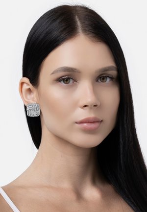 Cercei geometrice argintii cu un model împletit, având un finisaj mat. Purtați de un model cu păr drept închis la culoare și machiaj neutru.