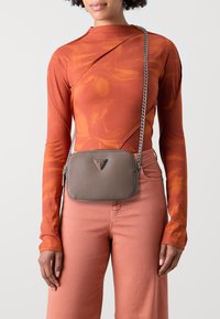 Borsa a tracolla marrone con finitura testurizzata, tracolla a catena argentata e logo triangolare. Indossata sopra un top a maniche lunghe arancione con motivo.