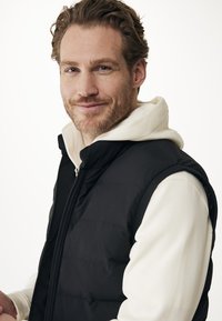 Muž se světle hnědými vlasy a vousy se usmívá, má na sobě bílé mikiny pod černou prošívanou vestou, na prostém světlém pozadí.