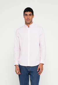 Camicia rosa chiaro con bottoni, maniche lunghe, colletto classico e una texture sottile, abbinata a jeans di denim scuro.