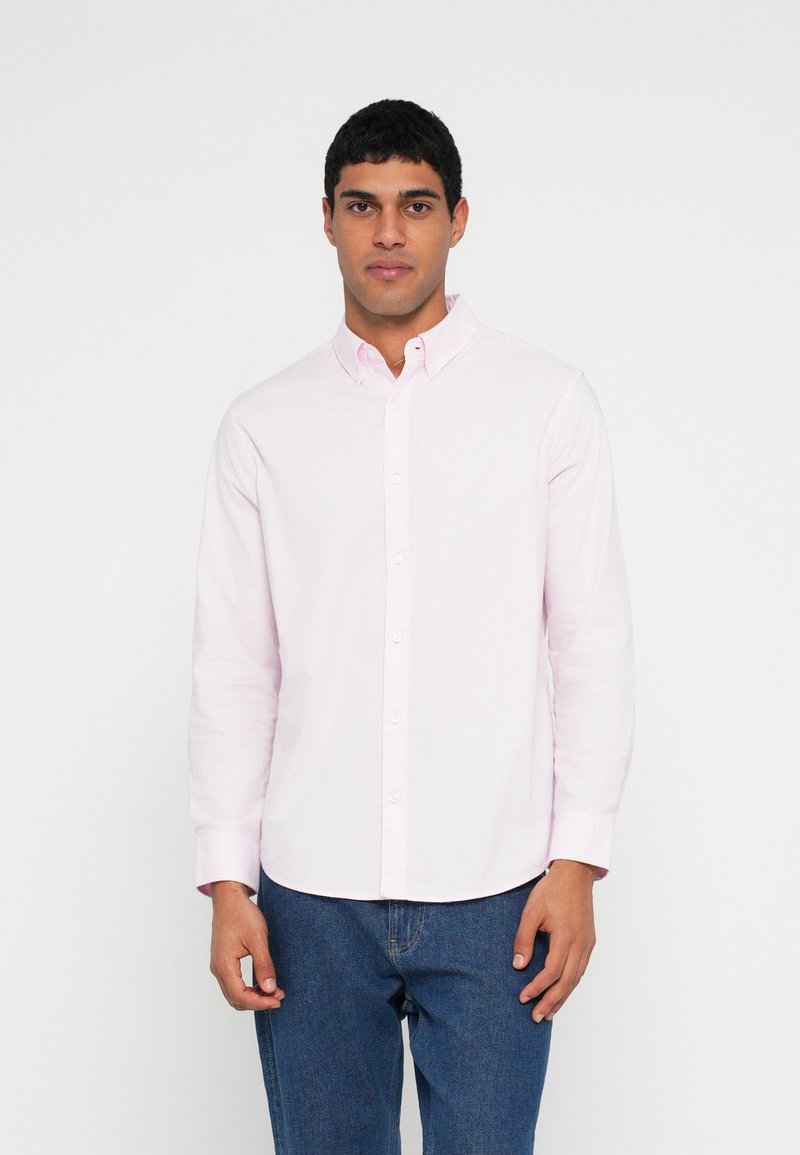 Camicia rosa chiaro con bottoni, maniche lunghe, colletto classico e una texture sottile, abbinata a jeans di denim scuro.