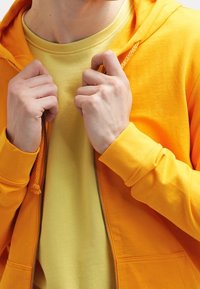 Sudadera amarilla con cremallera sobre una camiseta amarilla lisa. La sudadera tiene capucha con cordones y puños acanalados, manos sosteniendo la cremallera.