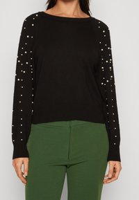 Schwarzer Strickpullover mit langen Ärmeln, verziert mit weißen Perlen entlang der Schultern, kombiniert mit enger grüner Hose.