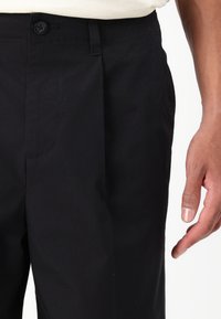 Black trousers with a straight cut, featuring a single button closure, and a smooth texture. Pants are partially tucked into a light shirt.