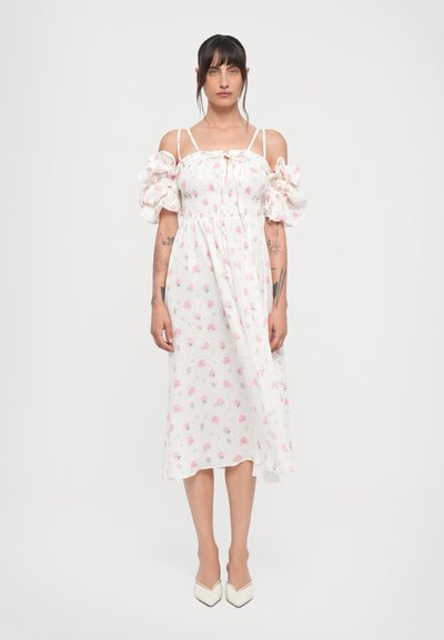 Hvid floral kjole med korte puffærmer, smocket liv og mønster af lyserøde roser. Båret med spidse hæle på en ensfarvet baggrund.
