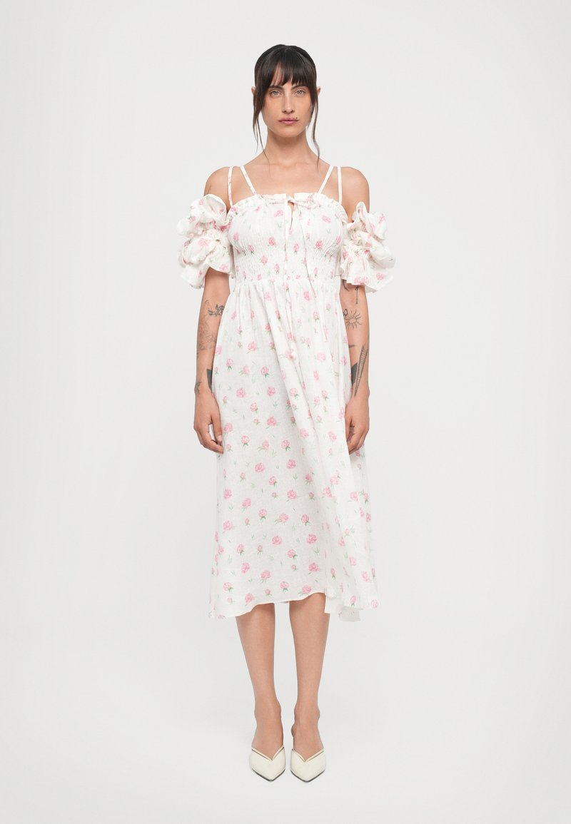 Hvid floral kjole med korte puffærmer, smocket liv og mønster af lyserøde roser. Båret med spidse hæle på en ensfarvet baggrund.