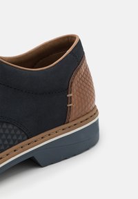 Chaussure en daim noir avec des accents texturés marron, présentant un talon à motif, une semelle grise et des coutures beiges sur le bord supérieur.