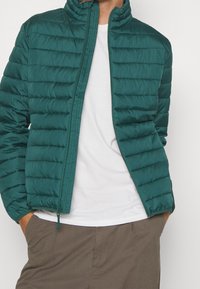 Teal puffer jacket, quilted design, high collar, front zipper, textured fabric, worn over a white shirt, paired with light brown pants.