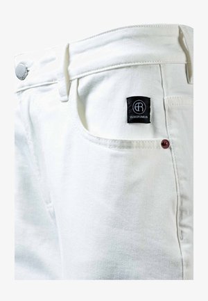 Primo piano di jeans in denim bianco che mostra la tasca frontale, il bottone d'argento e l'etichetta del marchio nera con il logo e il testo bianco "ELIASRUMELIS".