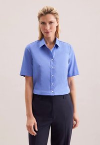 Light blue short-sleeve button-up shirt with a structured collar and front pleats, paired with dark tailored pants. Smooth fabric texture.