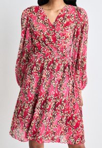 Robe portefeuille rouge à fleurs avec des manches longues, texture plissée et taille cintrée. Présente un motif de petites fleurs beiges et roses.