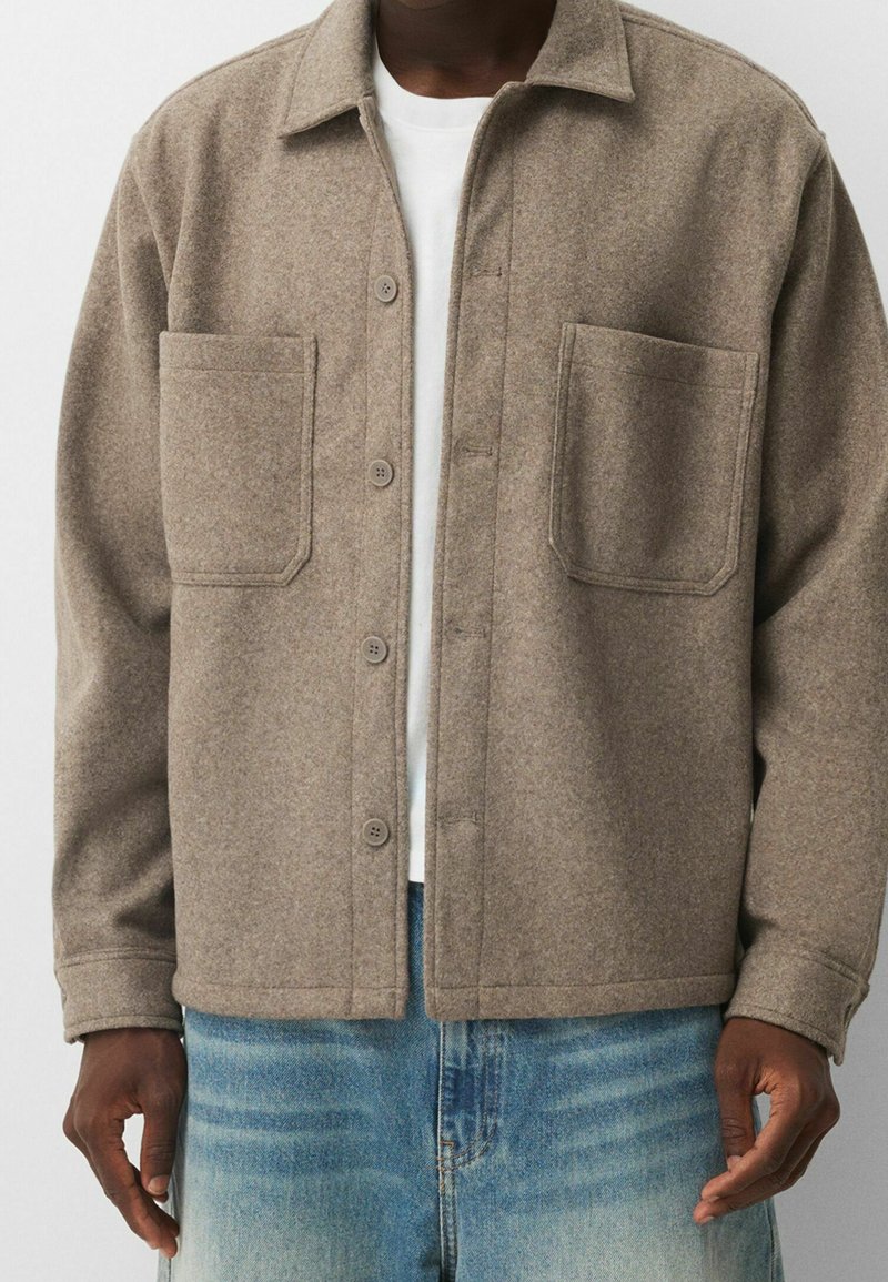 Homme portant une veste en laine beige à boutons avec deux poches avant, par-dessus une chemise blanche et un jean bleu, visible des épaules aux hanches.