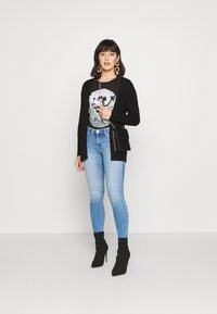 Svart cardigan over en grafisk t-skjorte, kombinert med lyseblå skinny jeans og svarte ankelboots med hæl, accessorert med en liten svart veske.