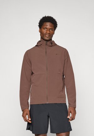 Veste légère marron avec une fermeture éclair à l'avant, capuche ajustable et poches. Texture douce, design minimal, avec un petit logo sur la poitrine.