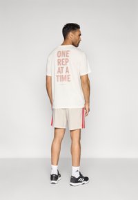 Ανοιχτόχρωμο t-shirt με τη φράση "ONE REP AT A TIME" σε έντονα κόκκινα γράμματα στην πλάτη, συνοδευόμενο από γκρι σορτς με κόκκινες ρίγες στα πλαϊνά. Μαύρα παπούτσια.