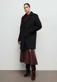 Czarny dwurzędowy płaszcz wełniany z szerokimi klapami, zestawiony z bordową plisowaną spódnicą i bordowymi kozakami za kolano, na neutralnym tle.