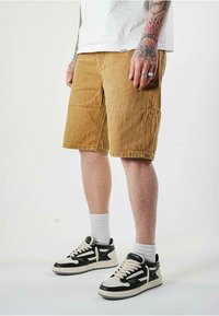 Pantaloni corti in corduroy color miele con taglio dritto e lunghezza al ginocchio. Indossati con calzini bianchi a coste e sneakers bicolore nere e bianche con strisce.
