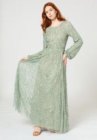 Lang groene jurk met ingewikkeld bead-patroon, doorschijnende stof, lange mouwen en een vloeiende rok. Gemaakt met open teen hakken.