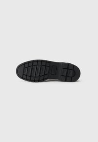 Semelle de chaussure en caoutchouc noir avec une texture, un motif antidérapant et plusieurs rainures de traction, affichant un design plat et une légère courbure.