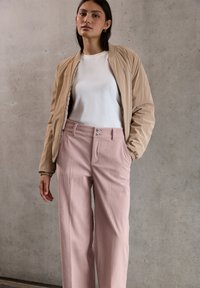 Donna in piedi contro un muro di cemento, indossa una giacca bomber beige sopra una maglietta bianca e pantaloni in corduroy rosa a vita alta, con le mani nelle tasche.