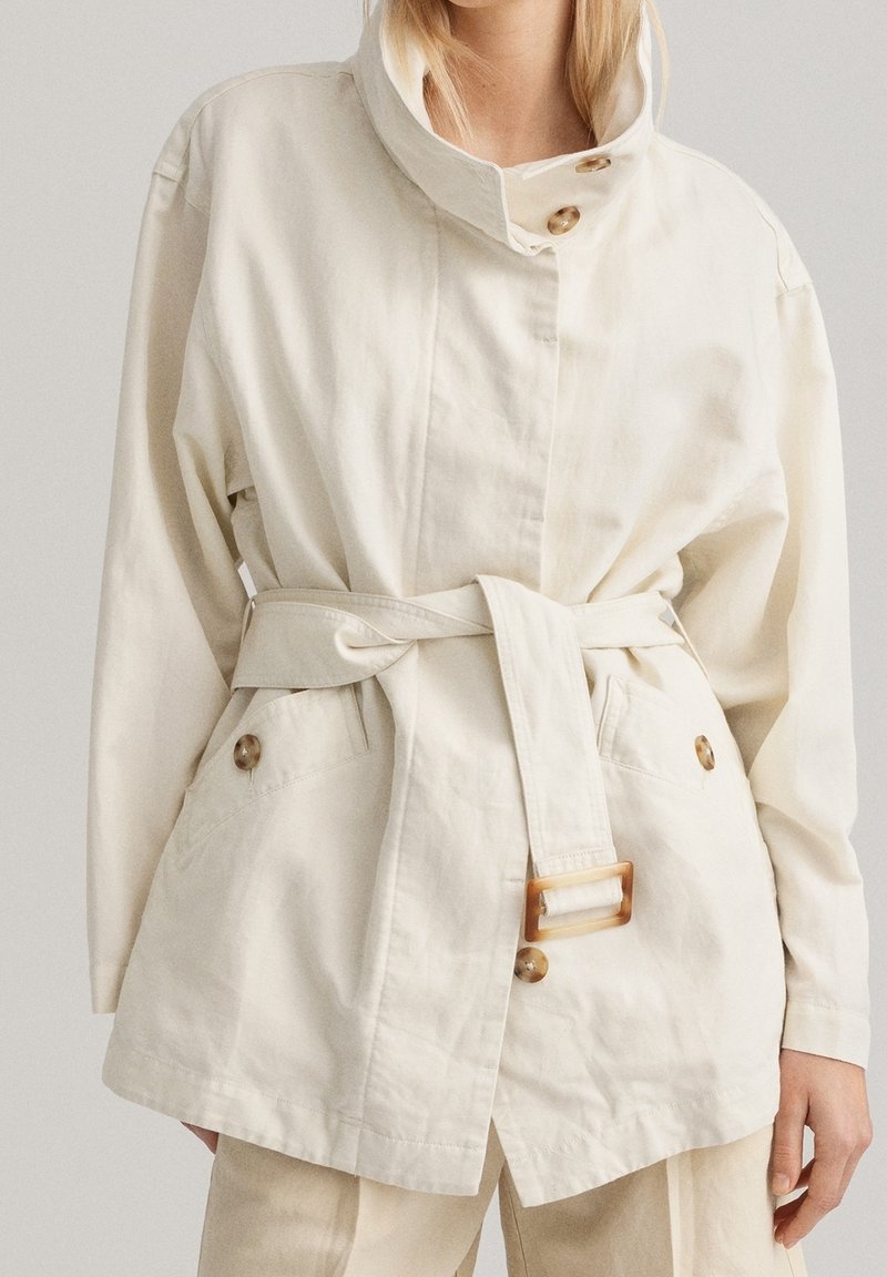 Giacca in cotone beige chiaro con colletto alto, chiusura a doppio bottone, vita con cintura e due tasche frontali. Presenta un tessuto liscio e testurizzato.