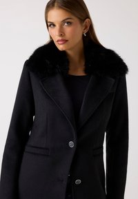 Sort uldfrakke med faux fur-krave, skræddersyet design, spidsede klapper og to frontlommer. Har sølvknapper til lukning.