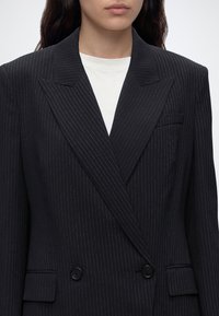 Femme portant un blazer noir à rayures fines, croisé, sur une chemise blanche, visible des épaules à la taille, debout devant un fond uni.