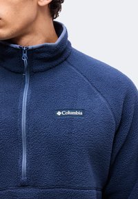 Άνδρας που φοράει μπλε σκούρο fleece μπουφάν Columbia με φερμουάρ μπροστά και ψηλό γιακά, κοιτάζοντας αριστερά, εμφανιζόμενος από το πηγούνι έως το στήθος.