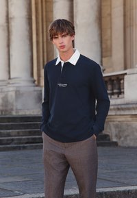 Tumesinine pika varrukaga polo valge kraega, millel on väike logo. Kandmisel pruunide ruuduliste pükstega. Taustal on kivist trepid ja sambad.