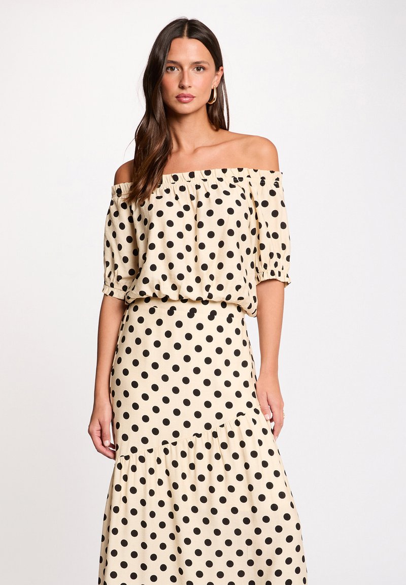 Mujer con blusa beige de palabra de honor y falda larga a juego con lunares negros, posando frente a un fondo blanco liso.