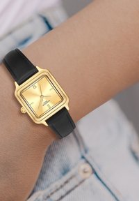 Montre carré avec un cadran doré et une finition mate, bracelet en cuir noir et marqueurs d'heure simples. Fixée à un poignet, avec un teint de peau clair visible.