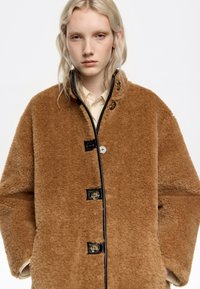 Joven con cabello rubio que lleva un abrigo de teddy marrón oversized con botones negros y una camisa beige, de pie contra un fondo blanco.