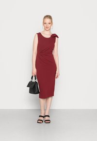 Vestido sem mangas bordô com detalhe em nó no ombro, silhueta ajustada, combinado com uma bolsa preta e sandálias pretas com tiras.