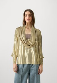 Top lungo a maniche con scollo drappeggiato in tessuto metallico oro, finitura testurizzata e vestibilità rilassata, abbinato a pantaloni di denim larghi.