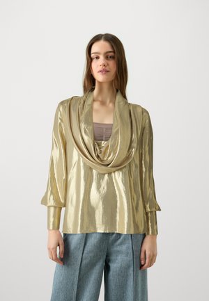 Gold metallic long-sleeve top with draped neckline, textured finish, and relaxed fit, paired with loose denim trousers.