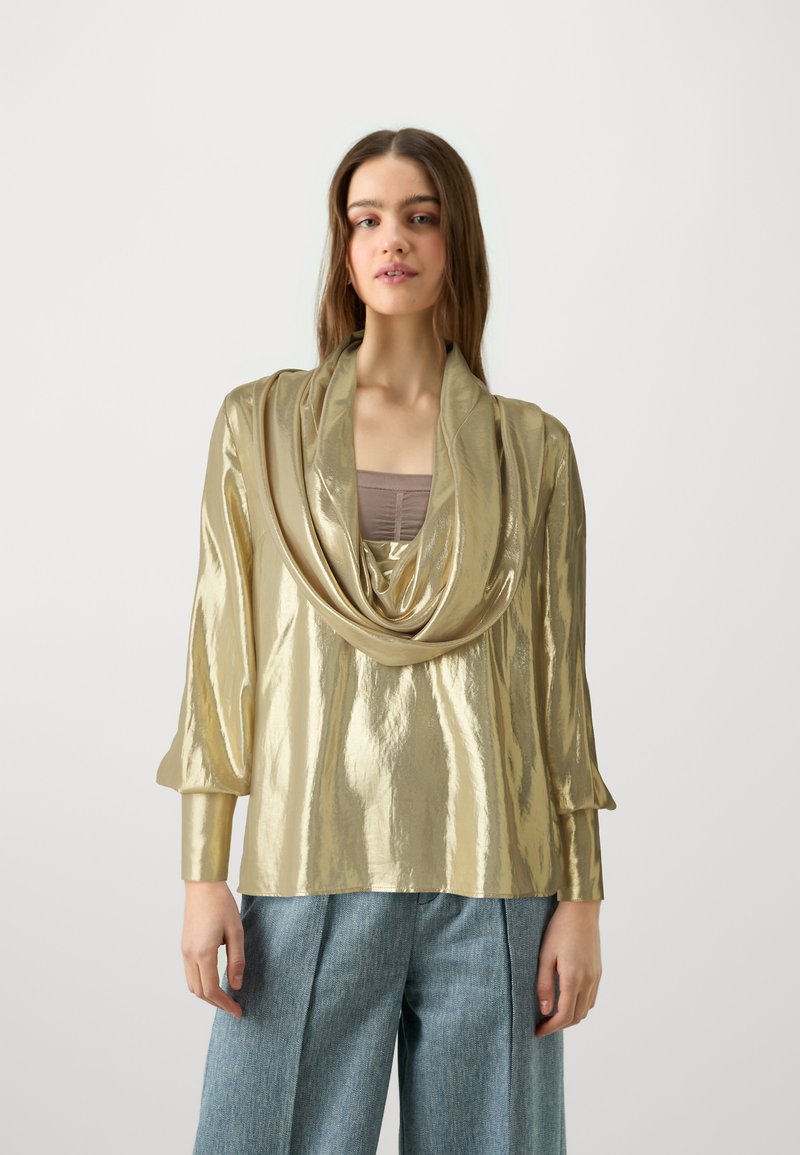 Top lungo a maniche con scollo drappeggiato in tessuto metallico oro, finitura testurizzata e vestibilità rilassata, abbinato a pantaloni di denim larghi.