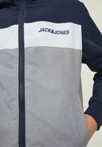 Marinblå, grå och vit zip-up jacka med en texturerad yta, med en logotyp på övre bröstet och sidofickor.