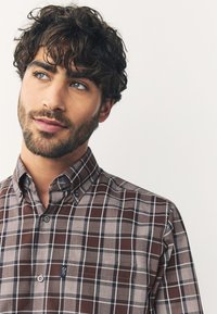 Chemise à carreaux marron à boutons avec col classique ; elle comporte une petite poche poitrine, des boutons foncés et des nuances variées de beige et de gris dans le motif.