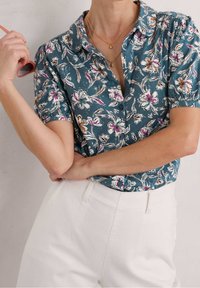 Camicia a maniche corte floreale in teal con accenti bianchi, rosa e marroni; indossata con pantaloni bianchi a vita alta.