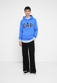Felpa con cappuccio blu con la scritta "GAP" in nero, indossata sopra una camicia a righe azzurra chiara, abbinata a pantaloni neri larghi e scarpe da ginnastica nere.