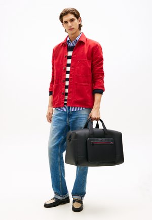 Borsa duffle nera con una tasca frontale con cerniera, texture liscia e manici robusti, abbinata a una camicia rossa e a un maglione a righe.