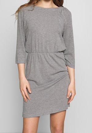 Femme portant une robe en maille gris clair, à manches longues, longueur genoux, avec une taille cintrée, sur un fond uni.