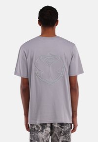 Grijze katoenen t-shirt met een groot geborduurd logo op de achterkant, met een oogontwerp binnen een schildvorm. Korte mouwen en een relaxte pasvorm.