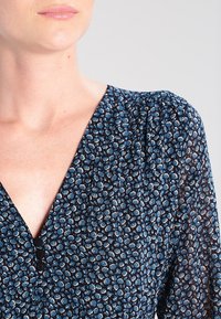 Niebieska sukienka w kwiatowy wzór z głębokim dekoltem w kształcie litery V, subtelnymi bufkami i małymi czarnymi guzikami przy dekolcie na ciemnym tle.