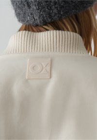 Personne portant une veste crème avec un col côtelé et un bonnet gris en tricot, montrant un écusson carré avec le logo sur la partie supérieure du dos de la veste.