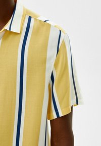 Camisa de manga corta en amarillo con rayas verticales en azul marino y blanco. Fabricada con un tejido ligero de textura suave y con cuello en punta.